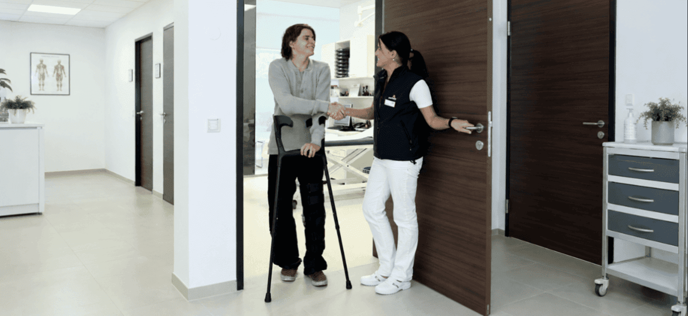 Vêem-se duas pessoas no corredor de uma clínica. Um homem com canadianas aperta a mão a uma médica.