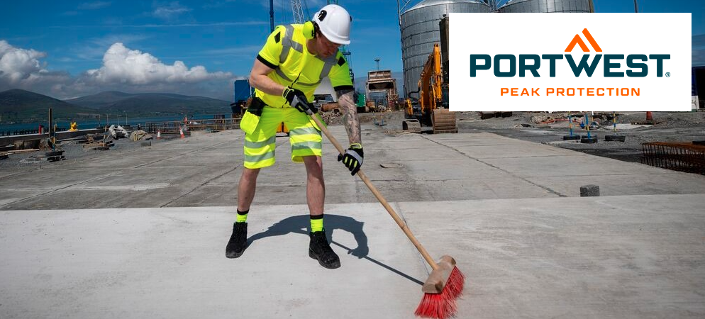 Um homem com um fato de trabalho amarelo de alta visibilidade e capacete branco está a varrer o chão de betão em frente a uma fábrica. Ao fundo, vêem-se montanhas e um céu azul com pequenas nuvens. O logótipo azul e laranja do fabricante, Portwest, está posicionado no canto superior direito.