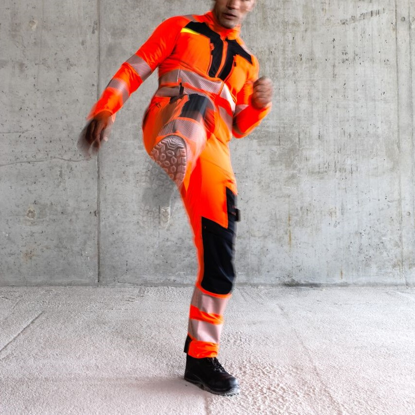 Trabalhador com roupa de prote&ccedil;&atilde;o laranja levanta uma perna num movimento de pontap&eacute;.