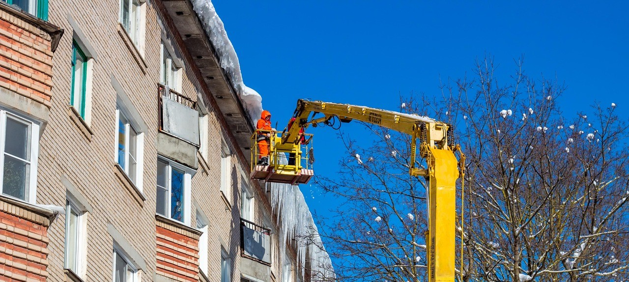 Uma fileira de casas com pingentes de gelo no telhado e neve sobre o mesmo. Um trabalhador com roupa laranja de alta visibilidade está numa plataforma elevatória. À direita, é visível uma árvore sem folhas com neve nos ramos.