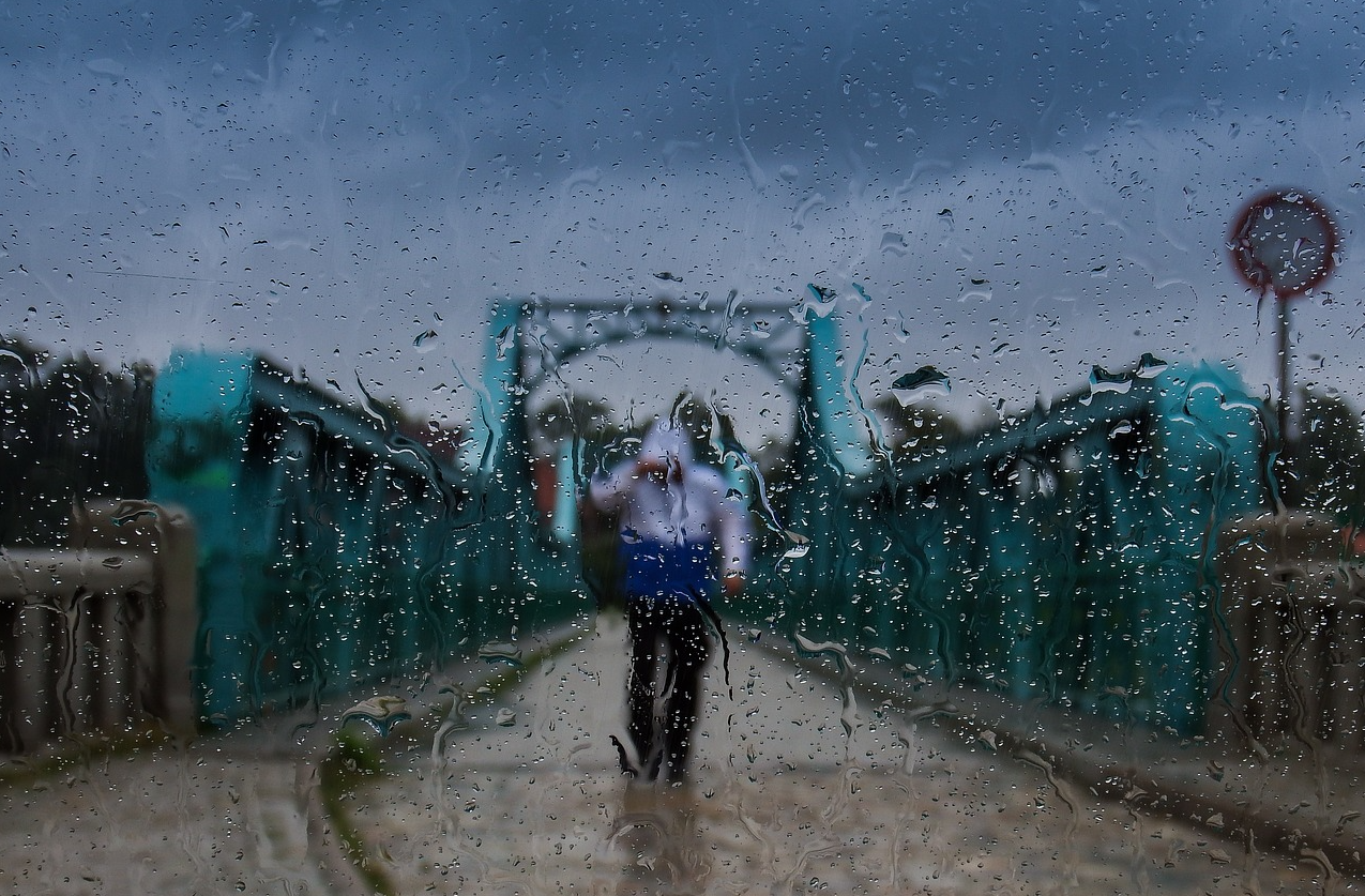Ponte azul contra um céu cinza com uma pessoa com capa de chuva no meio da estrada. A cena foi fotografada através de uma vidraça molhada. É fornecido um link para toda a nossa seleção de roupas de chuva práticas.