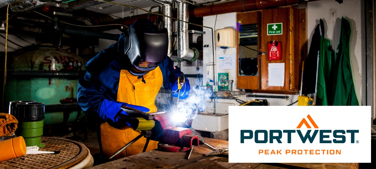 Um trabalhador usa uma máscara protetora e equipamento de proteção retardante de chamas em um ambiente industrial. Ele realiza trabalhos de soldagem e segura uma tocha de soldagem. Canos, ferramentas e um kit de primeiros socorros podem ser vistos ao fundo. No canto inferior direito da imagem está o logotipo “Portwest Peak Protection”.