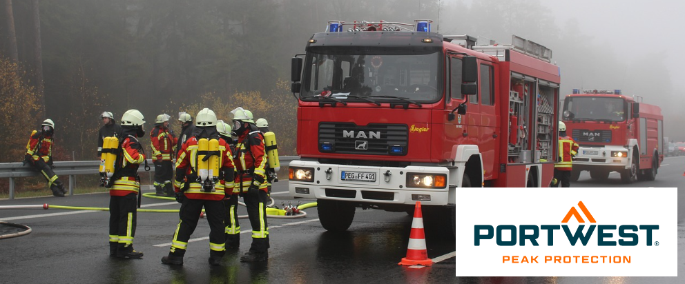 Cena de rua com dois carros de bombeiros vermelhos e vários bombeiros. Ao fundo você pode ver árvores cobertas de neblina. No canto inferior direito há o logotipo azul-laranja da Portwest na frente de um retângulo branco.