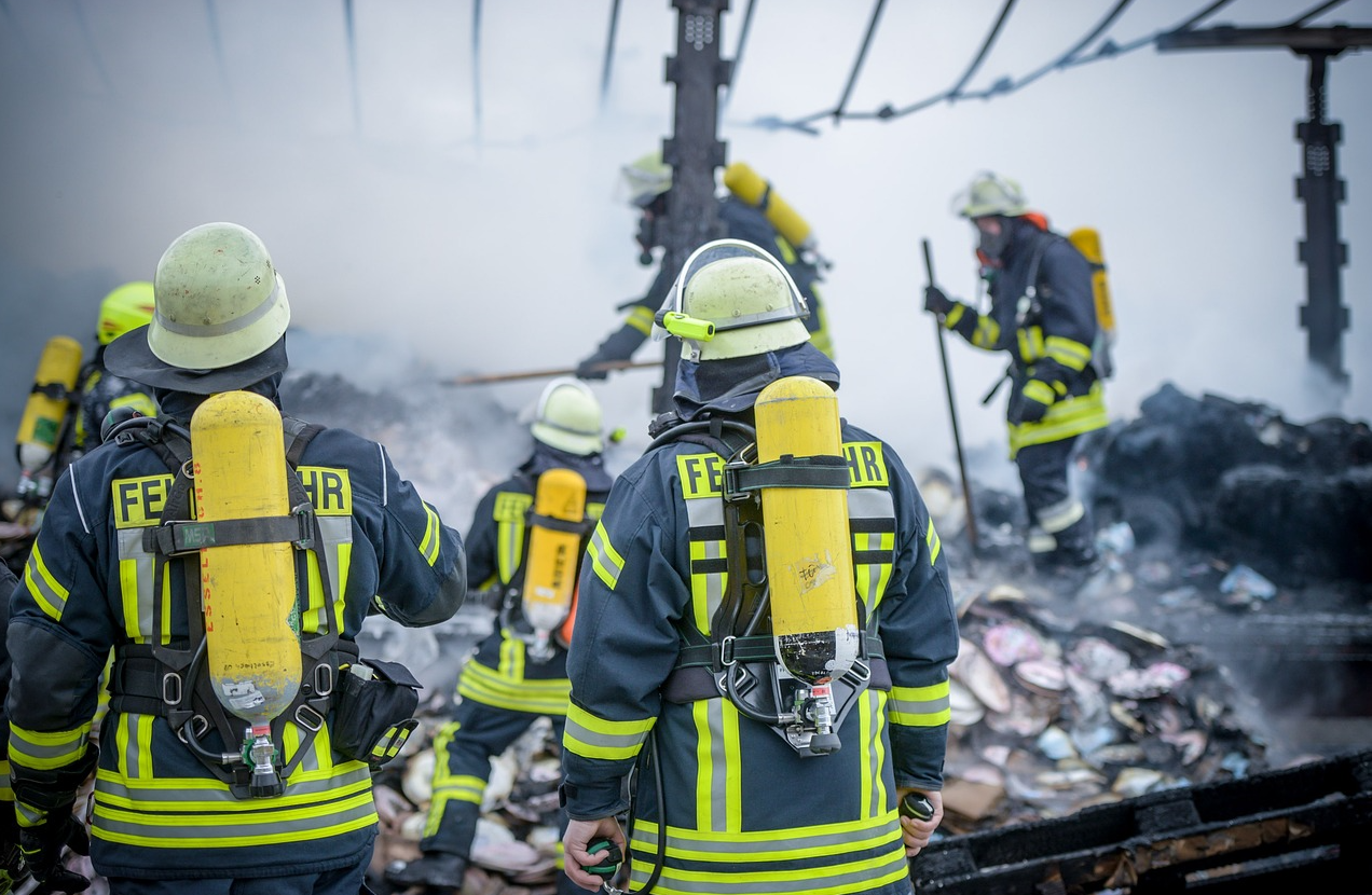Bombeiros em roupas azuis com listras amarelas de advertência e capacetes em um ferro-velho. O link fornecido leva à nossa seleção de roupas de combate a incêndios retardadoras de chamas.