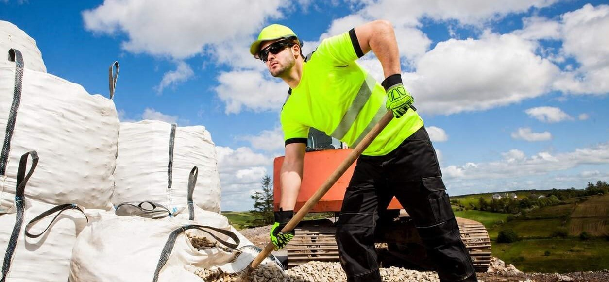 Um homem com camisola amarela e calças pretas. Usa um boné amarelo e óculos de sol e está a carregar cascalho de sacos grandes. Ao fundo, vê-se um céu azul com algumas nuvens, um estaleiro de obras e colinas verdes.