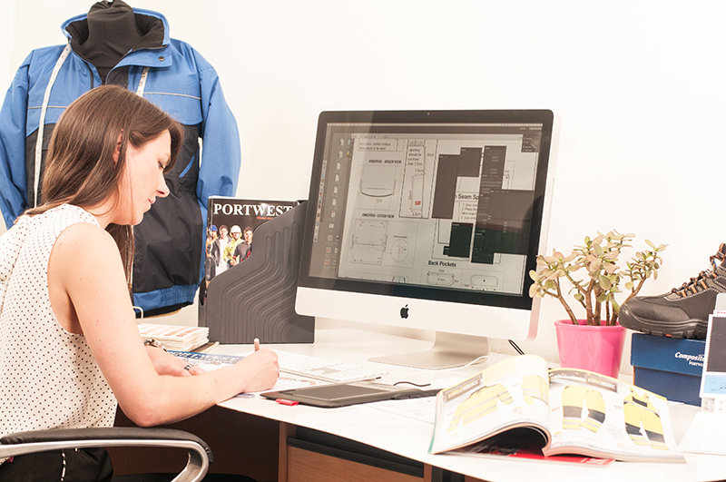 Mulher morena trabalhando na mesa. Desenhos técnicos de roupas de trabalho podem ser encontrados na tela do computador.