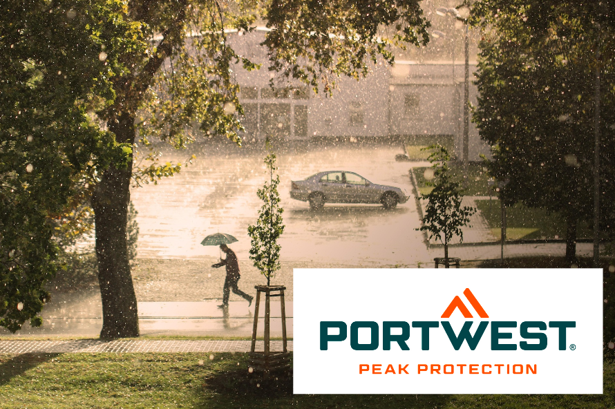 Vista de uma rua à chuva. Uma pessoa com um guarda-chuva atravessa a rua e o sol brilha mesmo chovendo. O logótipo do fabricante Portwest está localizado no canto inferior direito da imagem. Um link para as nossas capas de chuva é fornecido.