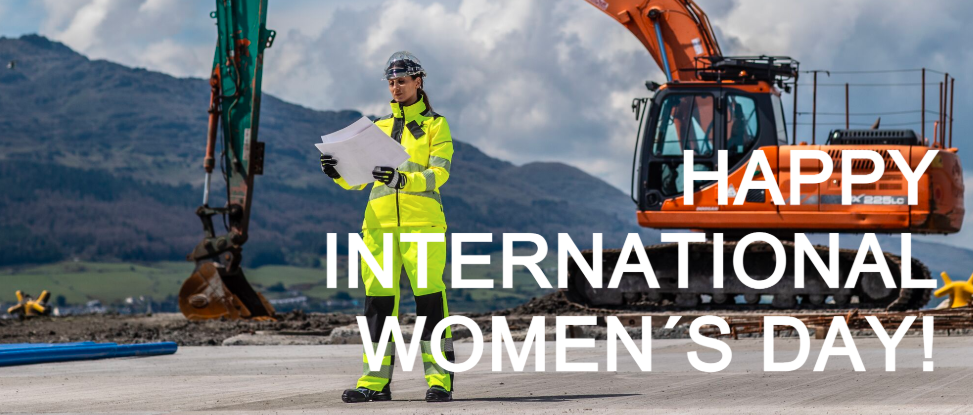 A imagem mostra uma mulher com equipamento de segurança amarelo brilhante, incluindo capacete e óculos de proteção, parada num estaleiro de construção a segurar plantas. Atrás dela, está uma grande escavadora laranja com o braço levantado e, ao fundo, colinas verdes estendem-se sob um céu parcialmente nublado. Por cima da imagem está escrito em grandes letras brancas "HAPPY INTERNATIONAL WOMEN´S DAY!" Em inglês.