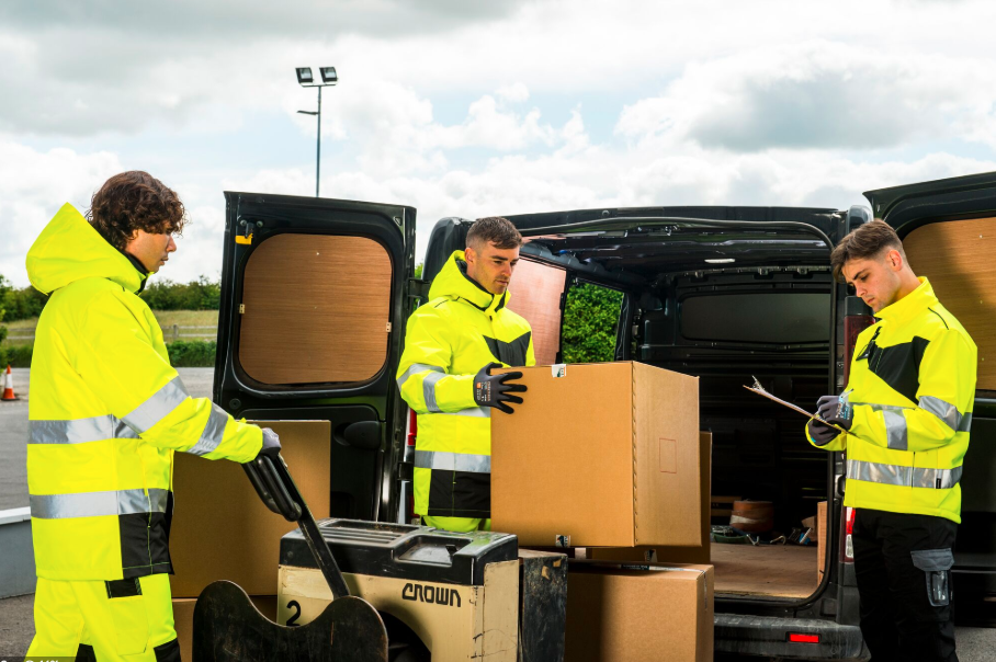 Três homens usando jaquetas softshell amarelo fluorescente com faixas refletivas trabalhando em frente a uma van aberta. Um empurra um palete de caixas com um transpalete, outro levanta uma grande caixa da van e o terceiro escreve algo em uma prancheta. A cena ocorre ao ar livre sob um céu nublado. Há um link para nossas jaquetas softshell.