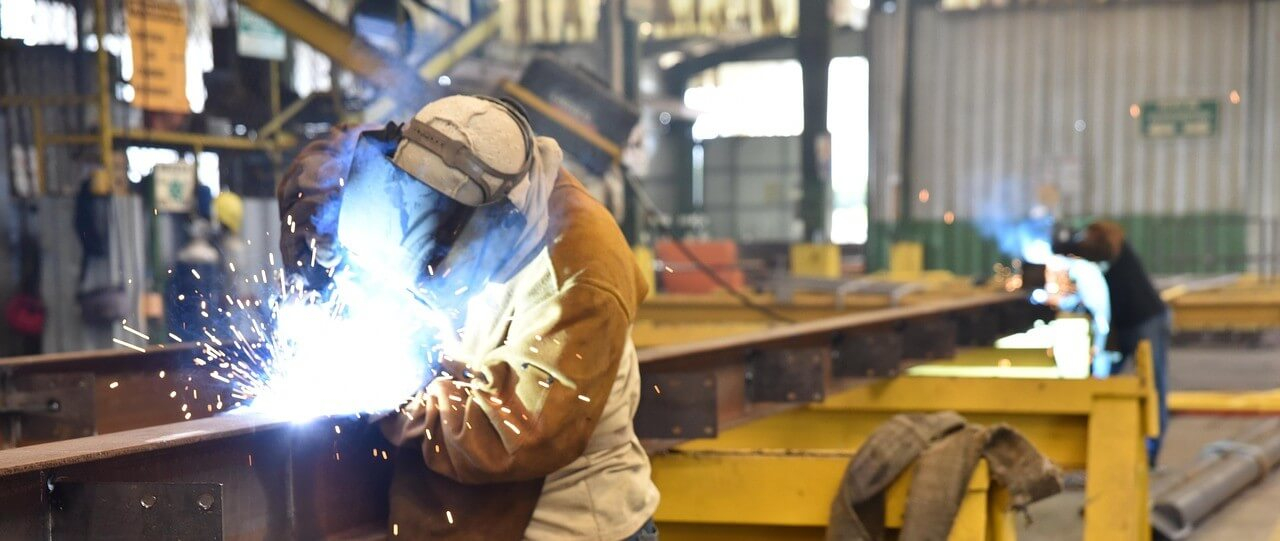 Vista de um barracão onde dois trabalhadores com equipamento de soldadura completo trabalham numa viga de aço.