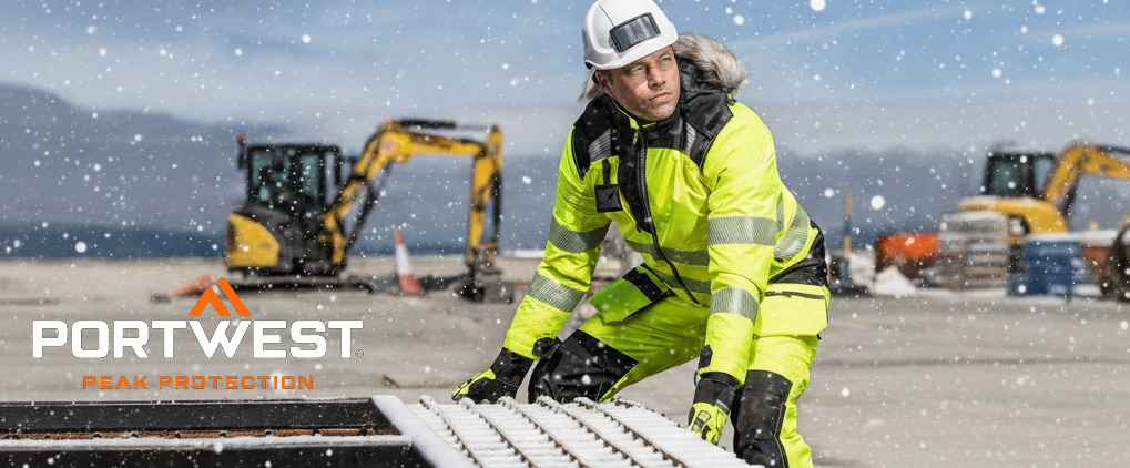 Um trabalhador com roupa de prote&ccedil;&atilde;o de inverno amarela e refletora trabalha ao ar livre sob neve. Usa capacete e luvas. Ve&iacute;culos de constru&ccedil;&atilde;o e uma paisagem nevada podem ser vistos ao fundo. No canto inferior esquerdo, o log&oacute;tipo do fabricante Portwest est&aacute; em letras brancas e laranja.