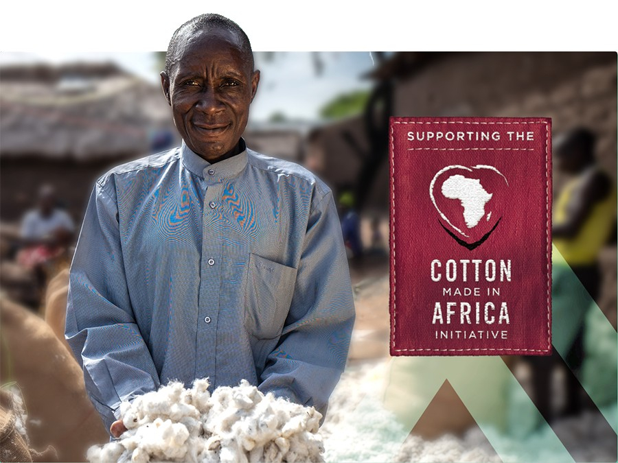 Agricultor africano vestindo uma camisa azul e apresentando um punhado de algodão. Uma placa vermelha com a inscrição "Apoiando a Iniciativa Cotton Made In Africa" ​​está na metade direita da imagem.