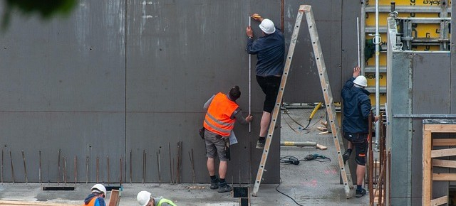 Dois trabalhadores são vistos num estaleiro de obras. Um está de pé numa escada, o outro segura-a. Ambos vestem calções de trabalho; um usa um capacete branco e o outro um colete de alta visibilidade laranja.