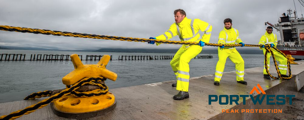 Três trabalhadores com roupa amarela de alta visibilidade a puxar uma corda. Ao fundo pode ver-se um navio, um porto e uma cadeia de montanhas nubladas, em primeiro plano está um poste amarelo. No canto inferior direito da imagem está o logótipo do fabricante Portwest em letras azuis e laranja.
