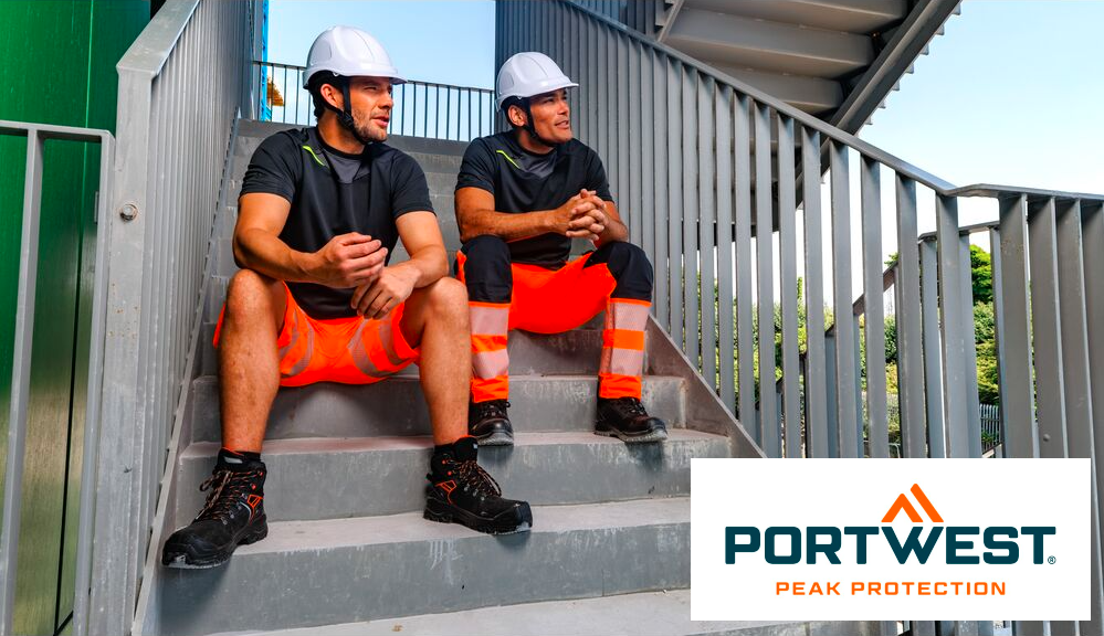 Dois trabalhadores com calças laranja de alta visibilidade e t-shirts pretas. Ambos os homens usam capacetes de segurança brancos e estão sentados numa escada com corrimão cinzento. Ao fundo pode ver o céu azul e as árvores, no canto inferior direito da imagem pode ver o logótipo do fabricante Portwest em letras azuis e laranja sobre um fundo branco. É fornecido um link para as nossas calças de trabalho.
