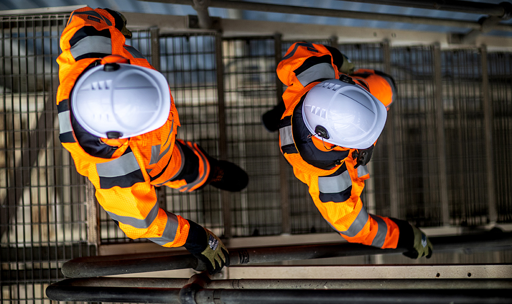 Visão aérea de dois trabalhadores usando capacetes brancos, luvas pretas e verdes, botas de segurança e macacões de alta visibilidade laranja com link para nossa coleção de roupas de proteção Arc Flash.