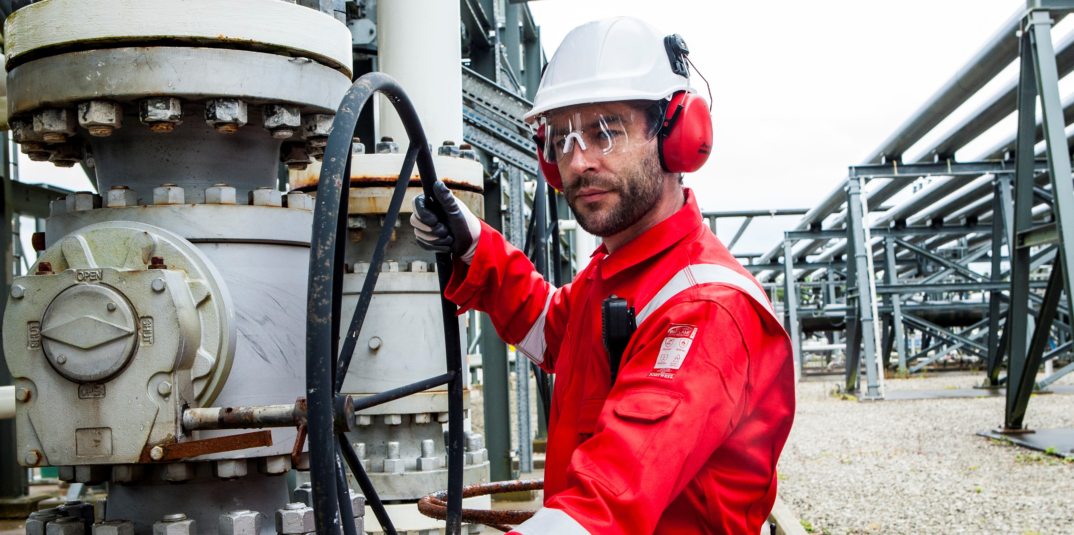 Trabalhador em contexto industrial com vestuário de proteção: capacete branco, óculos, orelheiras vermelhas, luvas pretas e fato-macaco FR50 vermelho com bandas refletoras.