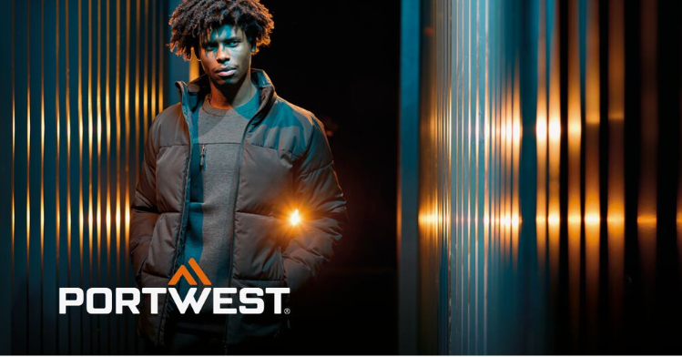 Um trabalhador com roupa de trabalho preta sobre um fundo abstrato com uma parede &agrave;s riscas e reflexos de luz. O log&oacute;tipo do fabricante, Portwest, em branco e laranja, est&aacute; localizado no canto inferior esquerdo da imagem.