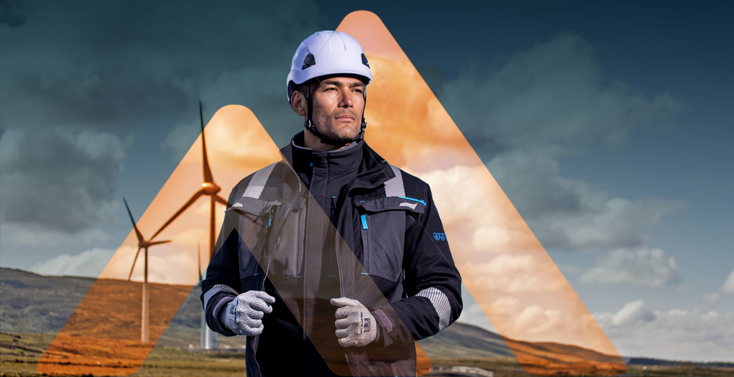 Homem a usar capacete, luvas e casaco de trabalho em frente a turbinas eólicas; ilustração de segurança no trabalho e proteção contra quedas em trabalhos em altura.