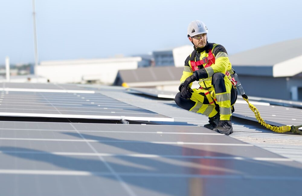 Trabalhador agachado num telhado entre instalações fotovoltaicas. Ele veste roupa de trabalho amarela de alta visibilidade, um capacete branco de proteção, luvas e sapatos pretos. Está seguro com o arnês antqueda Portwest 3-Ponto Plus FP18. O link leva à nossa categoria de proteção contra quedas.