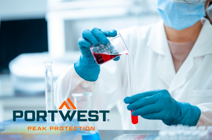 Trabalhador de laboratório que usa vestuário de proteção, bata, luvas azuis e máscara facial despeja líquido vermelho de um gobelé para um tubo de ensaio. Logótipo da Portwest com o slogan ‘Peak Protection’ em primeiro plano. É fornecido um link para a nossa seleção de batas.