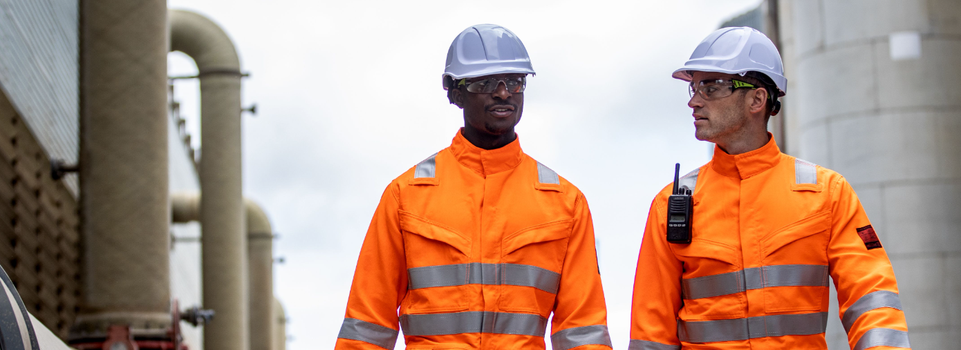 Dois trabalhadores em frente ao fundo industrial vestindo roupas laranja de alta visibilidade e capacetes brancos.