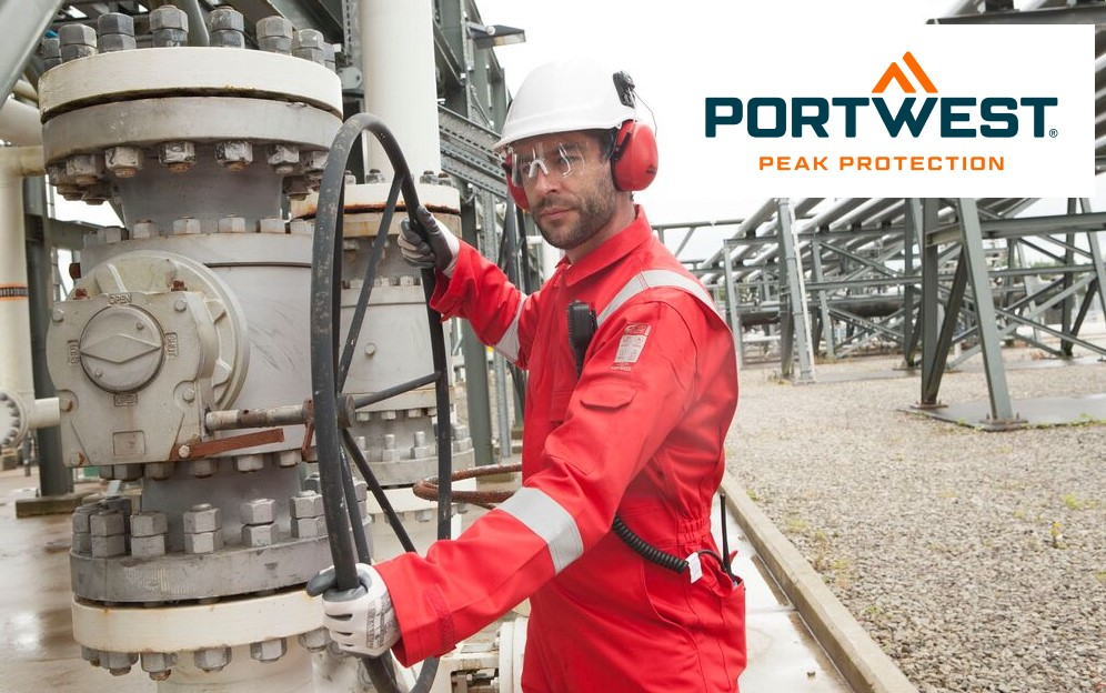 Um trabalhador industrial usa um fato de proteção vermelho com faixas refletoras, capacete branco, proteção auditiva e óculos de segurança. Opera um grande volante preto numa instalação industrial, presumivelmente numa refinaria ou pátio industrial. Tubagens e estruturas de aço podem ser vistas ao fundo. O logótipo da marca Portwest com o slogan "Peak Protection" está localizado no canto superior direito da imagem. Um link para os nossos fatos-macaco antichamas é fornecido.