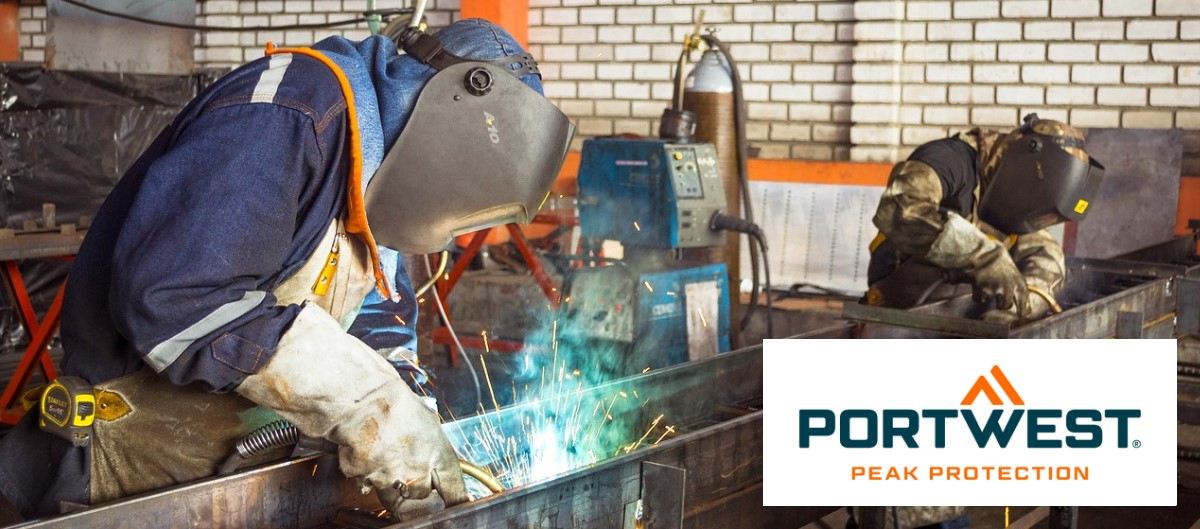 Vista de uma oficina de soldadura: dois trabalhadores com vestuário de proteção anti-chamas, luvas e máscaras de soldar estão a soldar uma longa chapa de metal.