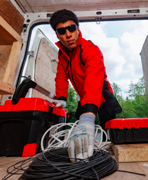 Um trabalhador que usa óculos escuros, casaco softshell vermelho, calças cargo pretas e luvas brancas alcança o interior de uma carrinha branca para apanhar um feixe de cabos pretos enrolados. Ao fundo, pode ver-se um céu azul com nuvens e árvores verdejantes. Um link leva à nossa seleção de luvas ESD.