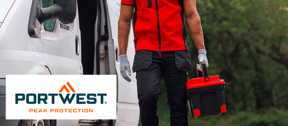 Pode ver a zona entre os ombros e os gémeos de uma pessoa com roupas de trabalho. O homem veste um colete vermelho softshell, uma t-shirt vermelha por baixo, calças pretas com bolsos e luvas brancas. Traz uma caixa de ferramentas na mão esquerda. À sua esquerda, ao fundo, pode ver uma carrinha branca; o restante fundo está coberto por árvores de folha caduca verdes. O logótipo azul e laranja do fabricante Portwest sobre fundo branco está localizado no canto inferior esquerdo da imagem.