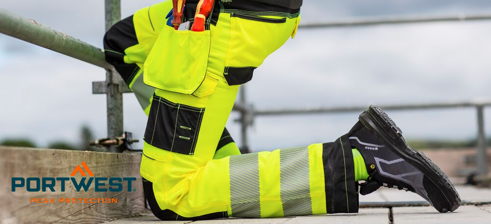 Um trabalhador da construção civil ajoelha-se no chão de um andaime. Usa calças de trabalho amarelas brilhantes de alta visibilidade com joelheiras integradas e calçado de segurança preto. Um céu nublado pode ser visto ao fundo. O logótipo do fabricante, Portwest, em letras azuis e laranja, está no canto inferior esquerdo.