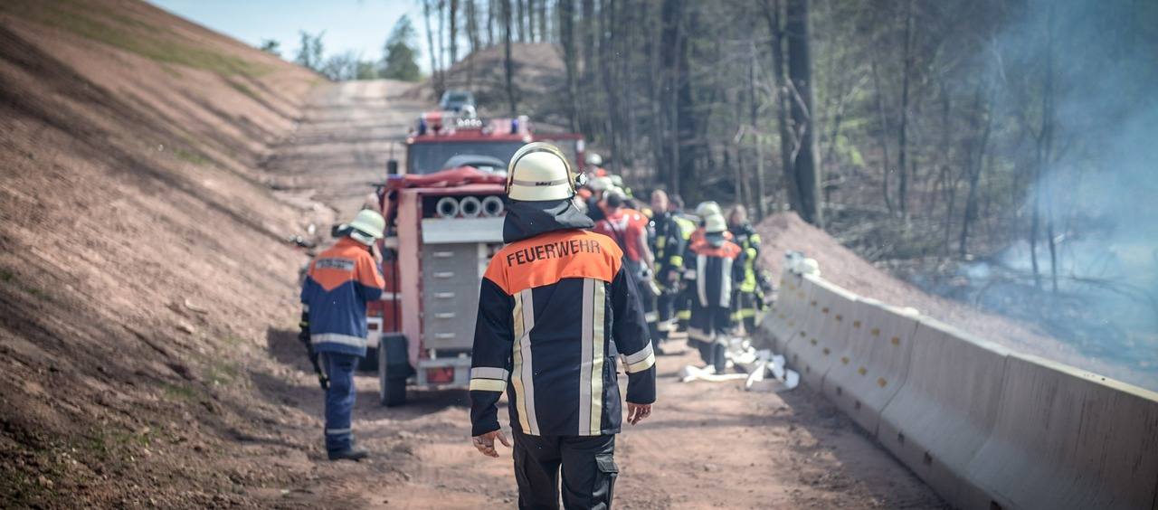 Vista traseira de um bombeiro em plena marcha. Ao fundo vê-se um carro de bombeiros num caminho florestal e um grupo de bombeiros a trabalhar. O link para o FR98 FR98 Bizflame FR98 FR98 Bizflame FR Fire Retardant Coverall é fornecido.