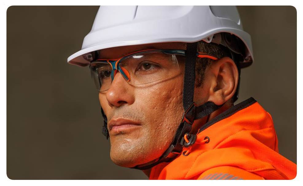 Retrato de um homem com vestuário de alta visibilidade laranja, capacete de proteção branco e óculos de segurança. Está incluída uma ligação para a nossa coleção de óculos.