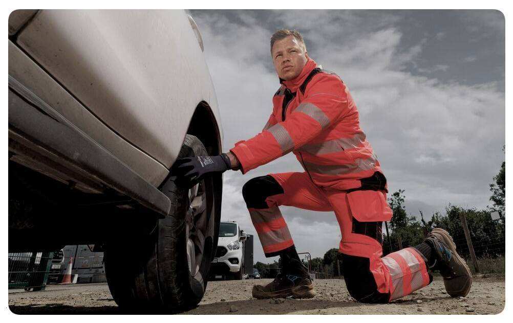 Um homem com vestuário de alta visibilidade laranja está ajoelhado em frente a um carro branco e trabalha no pneu. Ao fundo vê-se um céu nublado. Está incluída uma ligação para a nossa coleção de joelheiras.