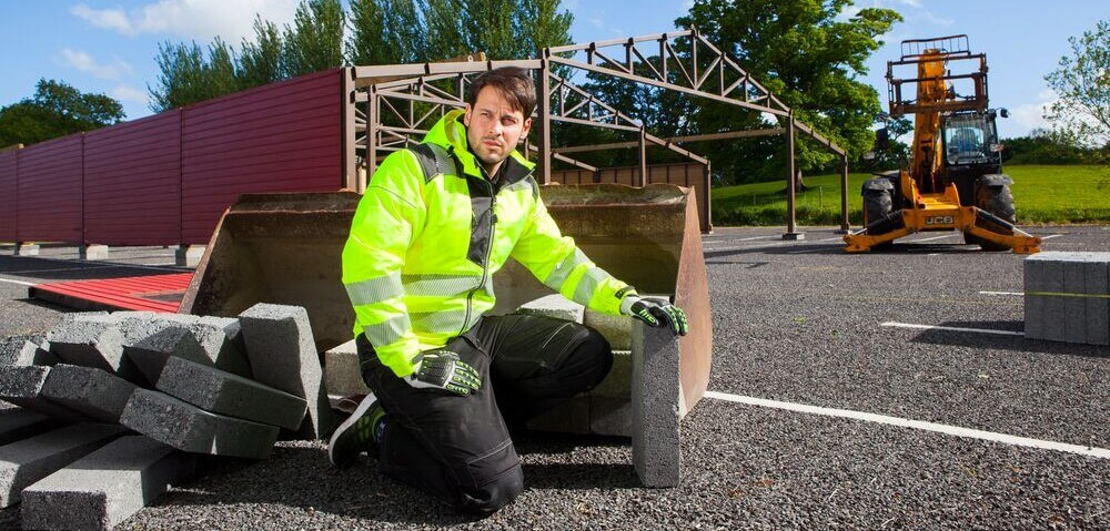 Um homem com um casaco de trabalho amarelo de alta visibilidade e calças pretas está ajoelhado na rua. Os materiais de construção e as máquinas são visíveis ao fundo.