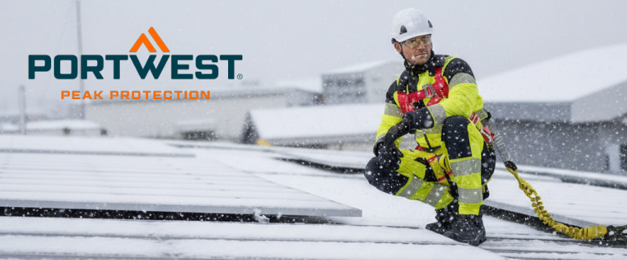 Um trabalhador com um fato-macaco amarelo de alta visibilidade com faixas refletoras ajoelha-se num telhado coberto de neve junto a um sistema fotovoltaico. Usa um capacete branco e equipamento de prote&ccedil;&atilde;o contra quedas. Mais telhados cobertos de neve s&atilde;o vis&iacute;veis em segundo plano, e o log&oacute;tipo do fabricante, Portwest, &eacute; vis&iacute;vel em azul e laranja no canto superior esquerdo da imagem.