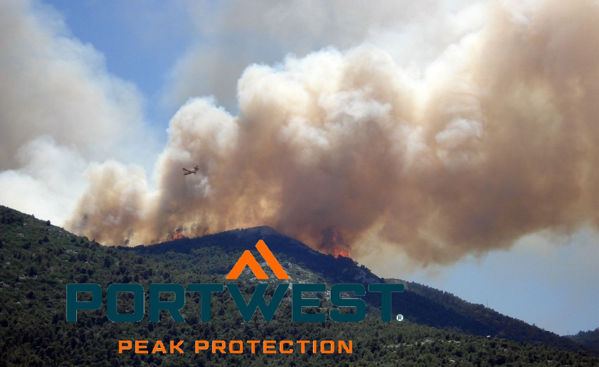 Um incêndio florestal assola uma encosta verdejante, com espessas nuvens de fumo a elevarem-se para o céu. Uma aeronave de combate a incêndios aproxima-se, despejando água ou agente extintor sobre as chamas. O logótipo da marca Portwest é visível em primeiro plano – o slogan "Peak Protection" está abaixo. Está incluído um link para a nossa seleção de vestuário de combate a incêndios.
